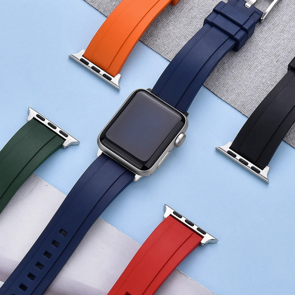 Top view of several rubber Apple Watch straps in different colors — red, navy blue (on watch), orange, green, and black — with silver adapters, displayed on a light blue background.